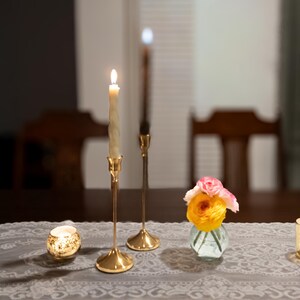 May include: A dimly lit table setting featuring two lit candles in gold candlesticks, a small vase of flowers, and two decorative candle holders. The table is covered with a gray lace tablecloth, creating a warm and inviting ambiance.