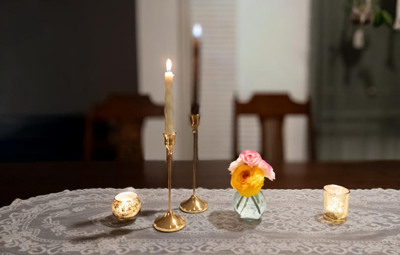 May include: A dimly lit table setting featuring two lit candles in gold candlesticks, a small vase of flowers, and two decorative candle holders. The table is covered with a gray lace tablecloth, creating a warm and inviting ambiance.