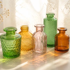 May include: A collection of five small glass vases in various colors and textures. The set includes a wide green vase with a honeycomb pattern, a yellow vase with a circular embossed design, a pink ribbed vase, a tall green vase, and a smooth amber vase.