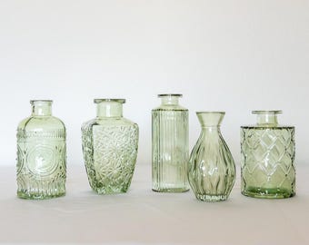 May include: Five decorative glass vases in varying shapes and designs. The vases are a pale green color and feature embossed patterns, vertical lines, and a smooth, rounded shape. They are arranged on a white surface.