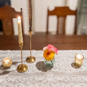 May include: A table setting featuring a white lace runner with gold candle holders, lit candles, and a small vase of flowers. The candles are lit, casting a warm glow on the table. The scene evokes a sense of elegance and tranquility.