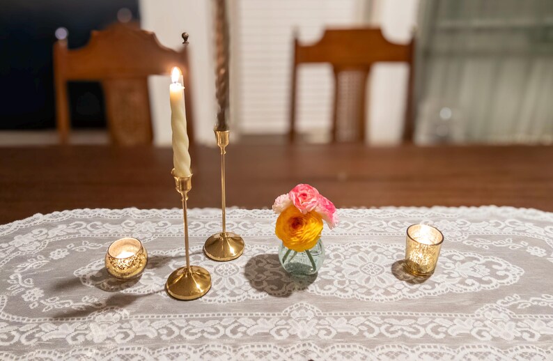 May include: A table setting featuring a white lace runner with gold candle holders, lit candles, and a small vase of flowers. The candles are lit, casting a warm glow on the table. The scene evokes a sense of elegance and tranquility.