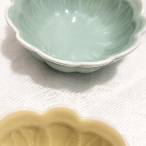 May include: Two ceramic bowls with scalloped edges. The top bowl is a pale green with a white rim, and the bottom bowl is a soft yellow. Both bowls are on a white, textured surface.