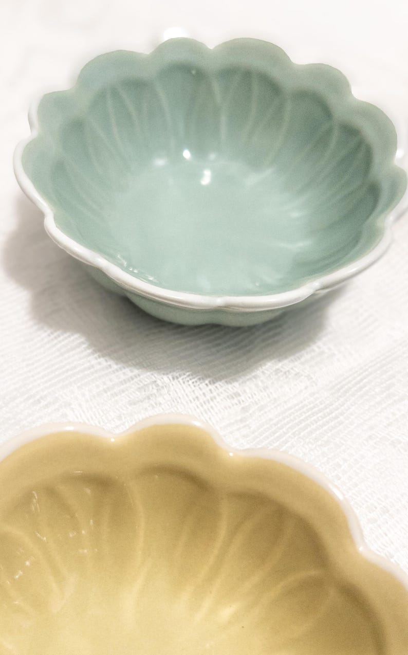 May include: Two ceramic bowls with scalloped edges. The top bowl is a pale green with a white rim, and the bottom bowl is a soft yellow. Both bowls are on a white, textured surface.