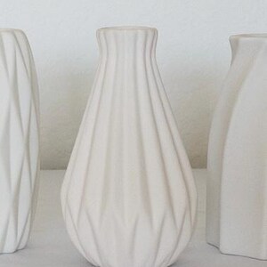Pu&ograve; includere: Tre vasi in ceramica bianca. Il vaso centrale ha un design strutturato e a coste. Gli altri due vasi hanno design geometrici e angolari. I vasi sono esposti su una superficie bianca su sfondo bianco.