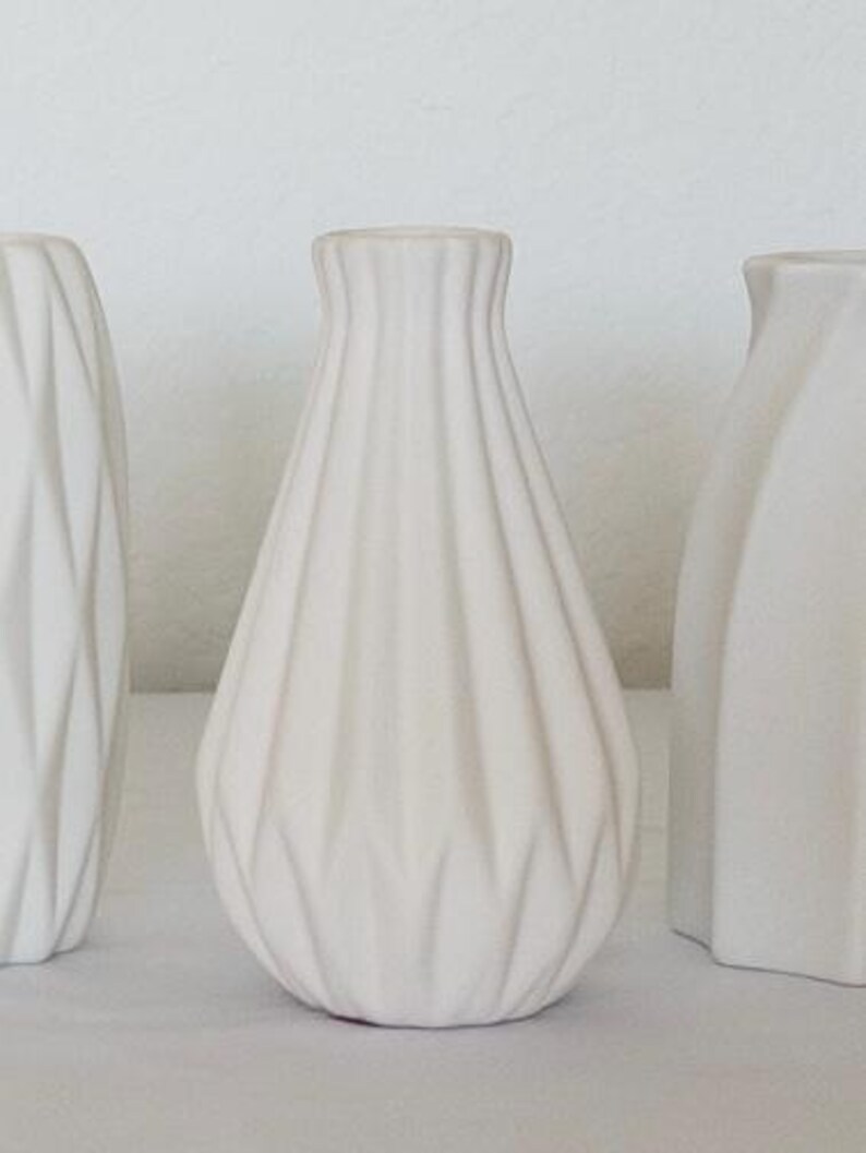 Pu&ograve; includere: Tre vasi in ceramica bianca. Il vaso centrale ha un design strutturato e a coste. Gli altri due vasi hanno design geometrici e angolari. I vasi sono esposti su una superficie bianca su sfondo bianco.