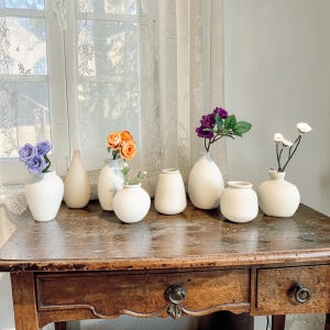 Könnte beinhalten: Eine weiße Keramik-Tasse mit schwarzem Henkel und dem Text "But First, Coffee" in fetter, schwarzer Schrift. Die Tasse steht vor einem verschwommenen Hintergrund, der eine gemütliche Umgebung suggeriert.