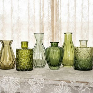 May include: A collection of six green glass vases in various shapes and sizes. The set includes vessels with geometric diamond patterns, vertical fluting, and smooth rounded silhouettes, all featuring translucent olive and emerald green glass.