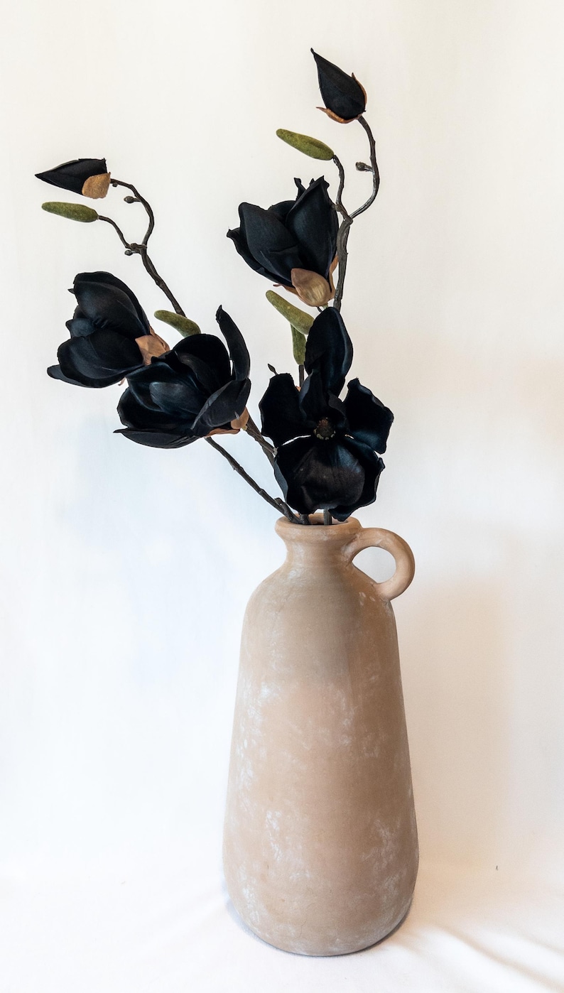 May include: A beige ceramic vase with a handle holds several black magnolia flowers and buds. The flowers have dark petals and are arranged with green and brown accents. The vase has a textured surface.