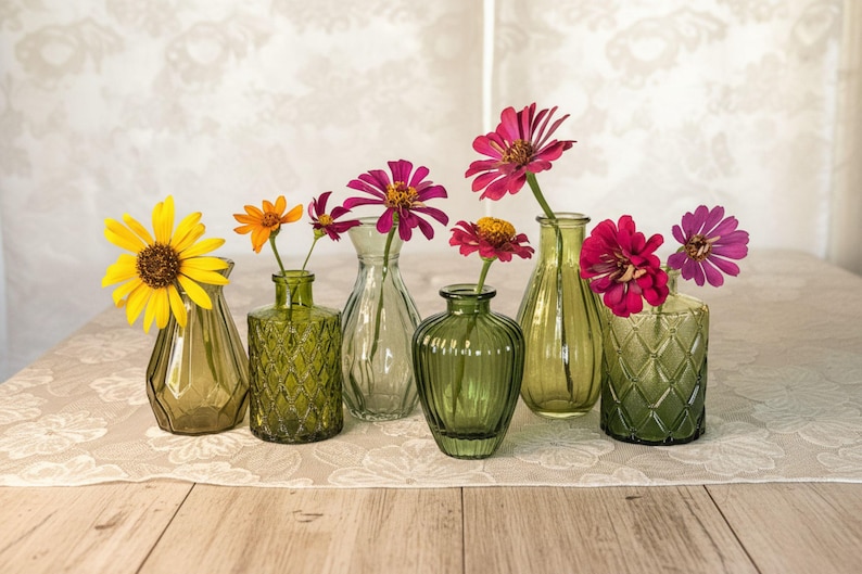 May include: A collection of six small green glass vases in various shapes and textures, each holding a single flower. The flowers include a large yellow daisy, orange blossoms, and several vibrant pink zinnia blooms with yellow centers.