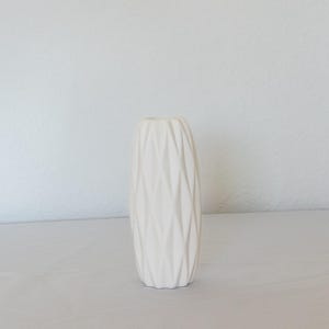 Pu&ograve; includere: Un vaso in ceramica color crema con un design geometrico strutturato. Il vaso &egrave; alto e cilindrico, con un collo stretto e una base larga. La superficie &egrave; ricoperta da un motivo ripetuto di linee angolate, creando un aspetto sfaccettato. Il vaso &egrave; vuoto e si trova su una superficie bianca.