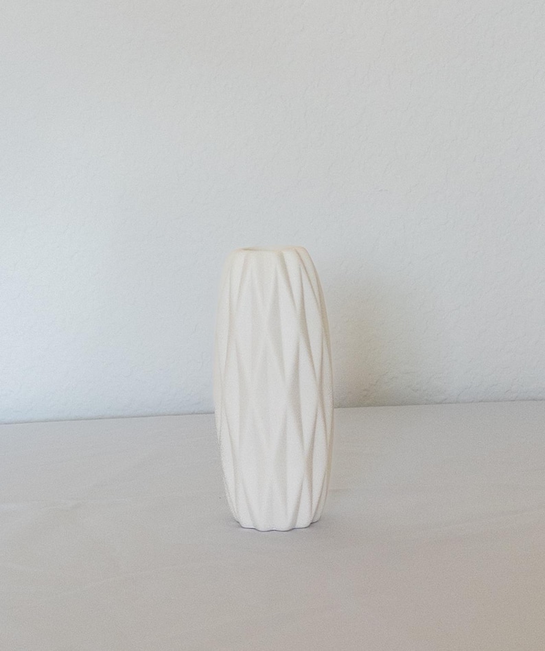 Pu&ograve; includere: Un vaso in ceramica color crema con un design geometrico strutturato. Il vaso &egrave; alto e cilindrico, con un collo stretto e una base larga. La superficie &egrave; ricoperta da un motivo ripetuto di linee angolate, creando un aspetto sfaccettato. Il vaso &egrave; vuoto e si trova su una superficie bianca.