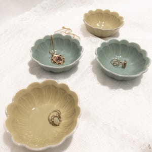 May include: Four small, scalloped-edge ceramic bowls in pale green and yellow hues. One bowl holds a gold necklace with a pendant, while another contains rings. The bowls sit on a white, textured surface, ideal for jewelry storage or display.