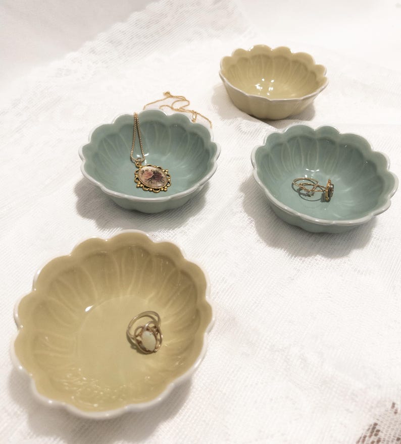 May include: Four small, scalloped-edge ceramic bowls in pale green and yellow hues. One bowl holds a gold necklace with a pendant, while another contains rings. The bowls sit on a white, textured surface, ideal for jewelry storage or display.