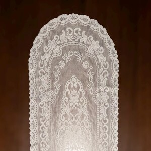 May include: White lace table runner with an intricate floral design. The runner has a scalloped edge and a symmetrical pattern of flowers and leaves. The delicate fabric is semi-transparent, revealing the surface beneath. This elegant runner is suitable for various home decor styles.