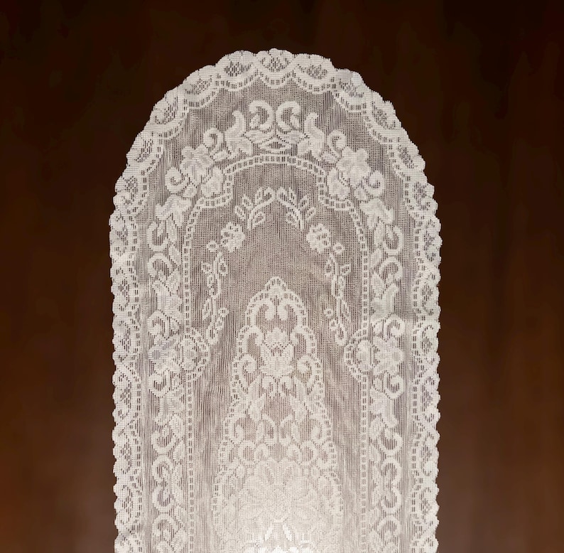 May include: White lace table runner with an intricate floral design. The runner has a scalloped edge and a symmetrical pattern of flowers and leaves. The delicate fabric is semi-transparent, revealing the surface beneath. This elegant runner is suitable for various home decor styles.