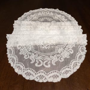 May include: A stack of white lace doilies with intricate floral and scrollwork patterns. The doilies have a scalloped edge and are made of a delicate, sheer fabric. The doilies are arranged on a dark brown surface.