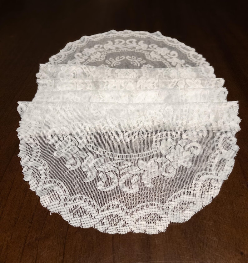 May include: A stack of white lace doilies with intricate floral and scrollwork patterns. The doilies have a scalloped edge and are made of a delicate, sheer fabric. The doilies are arranged on a dark brown surface.