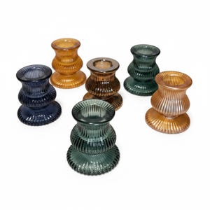 Könnte beinhalten: Eine Sammlung von sechs Glaskerzenhaltern in verschiedenen Farben, darunter Blau, Bernstein, Braun, Grün und Hellbraun. Jeder Halter hat ein abgerundetes, geripptes und geriffeltes Design mit einem breiten Fuß und einer runden Öffnung oben.