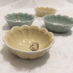 May include: A collection of small, scalloped-edge ceramic bowls in pastel yellow and green hues. One bowl holds a silver ring with an oval, off-white stone. The bowls are arranged on a white, textured surface, suggesting a decorative or storage theme.