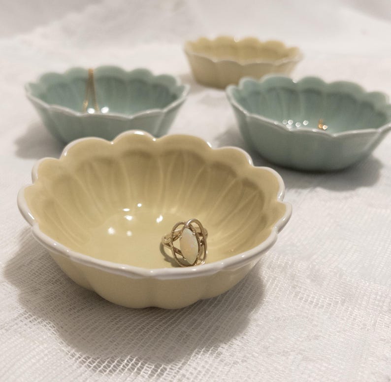 May include: A collection of small, scalloped-edge ceramic bowls in pastel yellow and green hues. One bowl holds a silver ring with an oval, off-white stone. The bowls are arranged on a white, textured surface, suggesting a decorative or storage theme.