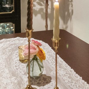 May include: A table setting with lit candles and floral arrangements. Two gold candlesticks hold lit candles, one brown and twisted, the other cream-colored. A small vase with pink and yellow flowers sits between them.