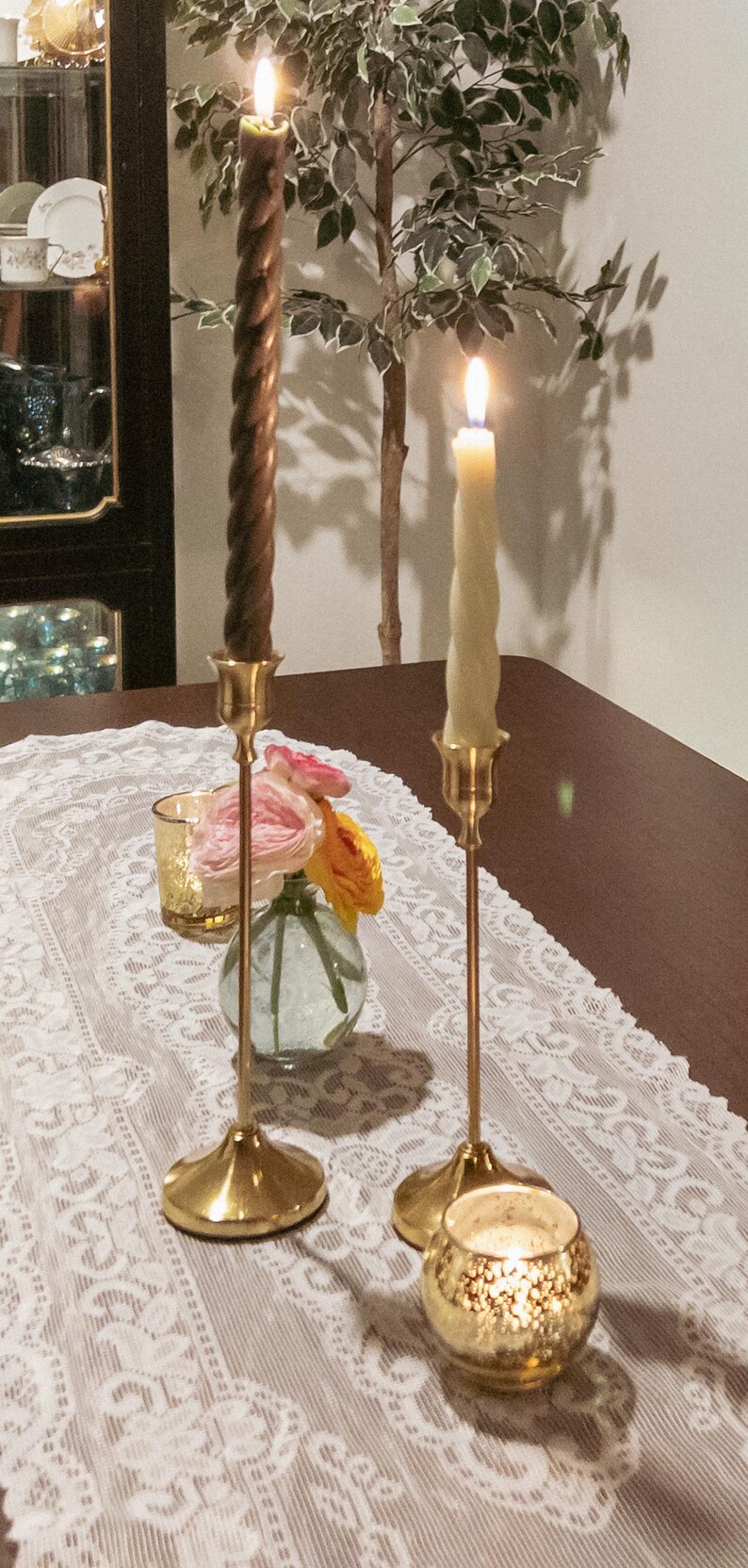 May include: A table setting with lit candles and floral arrangements. Two gold candlesticks hold lit candles, one brown and twisted, the other cream-colored. A small vase with pink and yellow flowers sits between them.