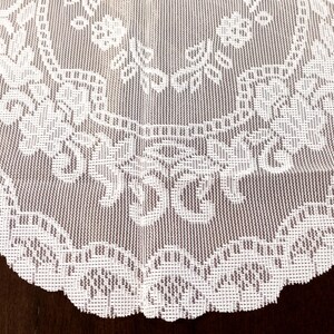 May include: A close-up of a white lace doily with a floral pattern. The doily has a scalloped edge and is made of a delicate, open-weave fabric. The intricate design features flowers and leaves.