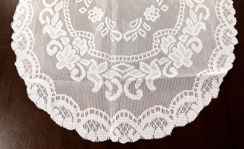 May include: A close-up of a white lace doily with a floral pattern. The doily has a scalloped edge and is made of a delicate, open-weave fabric. The intricate design features flowers and leaves.