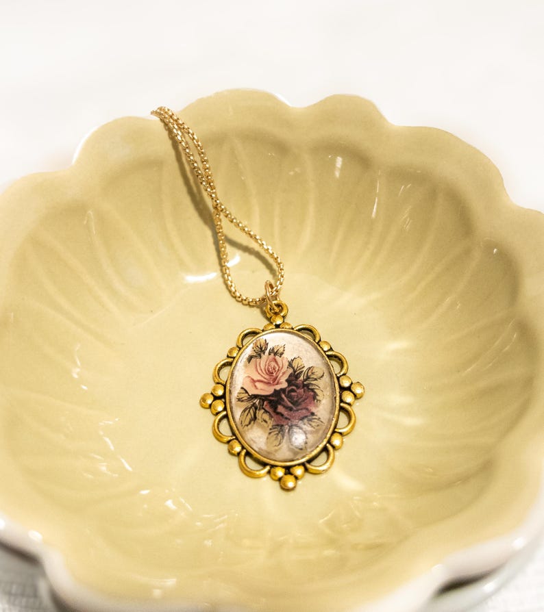May include: A gold-colored pendant necklace featuring an oval-shaped pendant with a floral design. The pendant showcases a pink and burgundy rose bouquet. The necklace is set against a light yellow, scalloped dish.