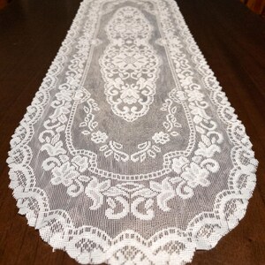 May include: White lace table runner with an intricate floral design. The runner is rectangular with a scalloped edge. The delicate lace pattern is visible throughout the entire piece, adding a touch of elegance to any table setting.