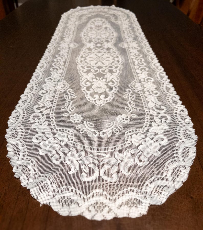 May include: White lace table runner with an intricate floral design. The runner is rectangular with a scalloped edge. The delicate lace pattern is visible throughout the entire piece, adding a touch of elegance to any table setting.