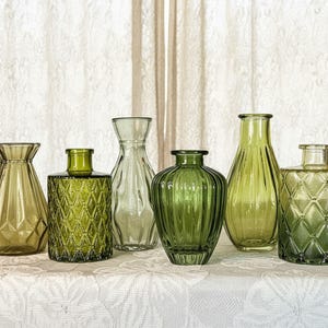 May include: A collection of six glass vases in various shades of green. The set includes vessels with geometric diamond patterns, vertical ribbed textures, and smooth tapered silhouettes, all featuring narrow openings and transparent glass construction.