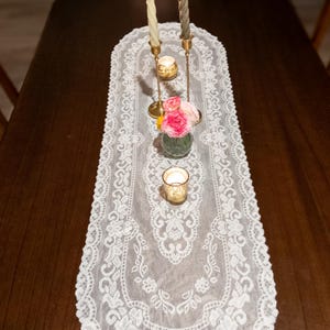 May include: A white lace table runner with a floral design, adorned with candles and a small vase of pink roses. Two lit candles stand tall, and a small votive candle holder is also present. The table is made of dark wood.