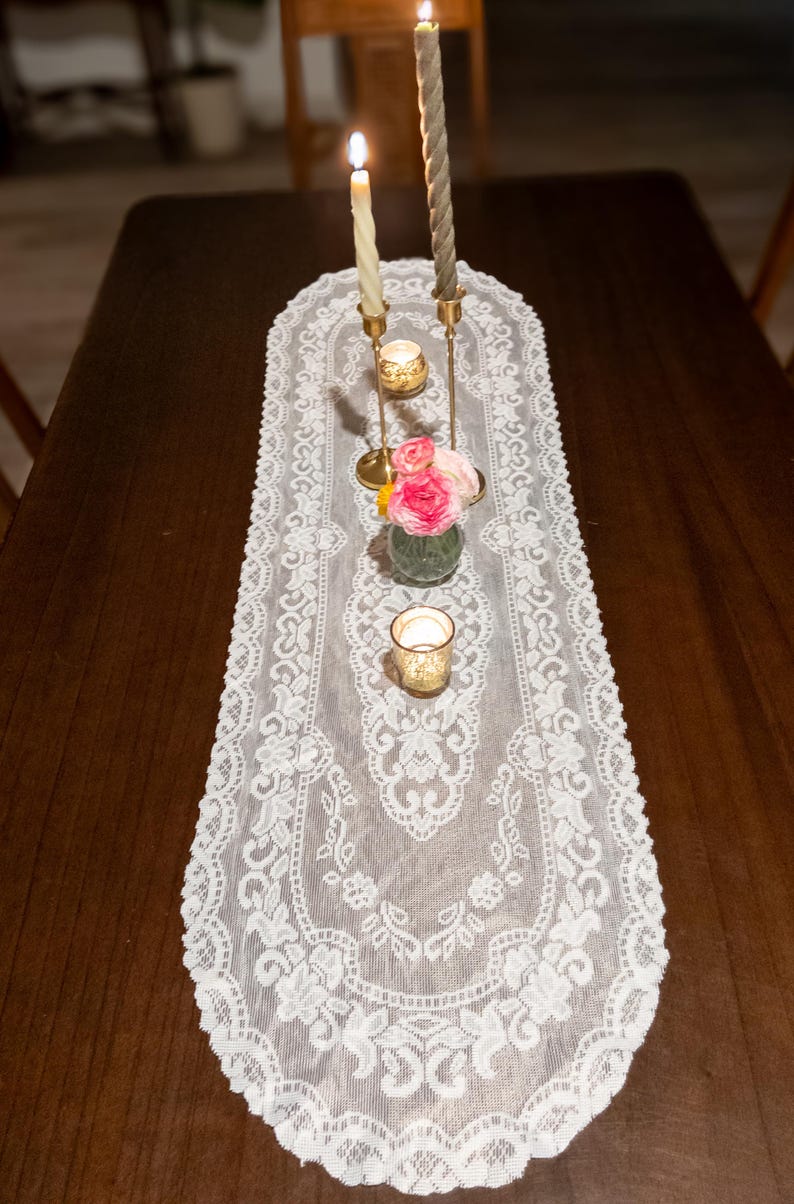 May include: A white lace table runner with a floral design, adorned with candles and a small vase of pink roses. Two lit candles stand tall, and a small votive candle holder is also present. The table is made of dark wood.