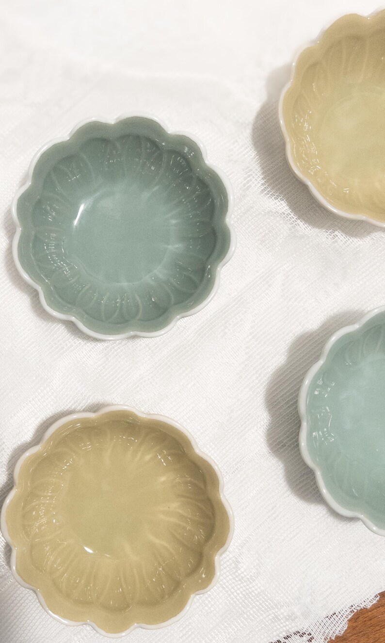 May include: Four small, decorative ceramic bowls with a scalloped edge and a floral pattern. Two bowls are a pale green, and two are a pale yellow. The bowls are on a white, textured surface.