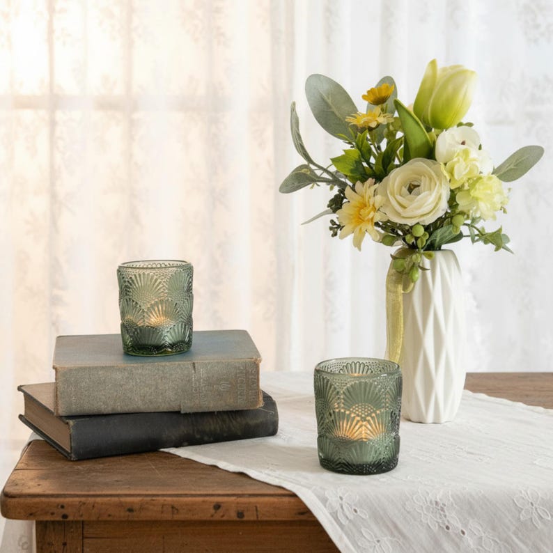 K&ouml;nnte beinhalten: Zwei salbeigr&uuml;ne Glas-Teelichthalter mit Muschelmuster, einer auf zwei gestapelten alten B&uuml;chern und der andere auf einem wei&szlig;en Tischl&auml;ufer. Eine wei&szlig;e geometrische Vase enth&auml;lt einen Strau&szlig; aus cremefarbenen und gelben Blumen mit gr&uuml;nem Blattwerk.