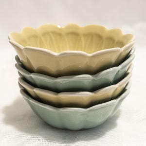 May include: A stack of four ceramic bowls in pastel colors. The bowls have a scalloped edge and are in shades of yellow and green. The bowls are stacked on top of each other.
