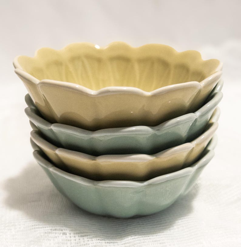 May include: A stack of four ceramic bowls in pastel colors. The bowls have a scalloped edge and are in shades of yellow and green. The bowls are stacked on top of each other.
