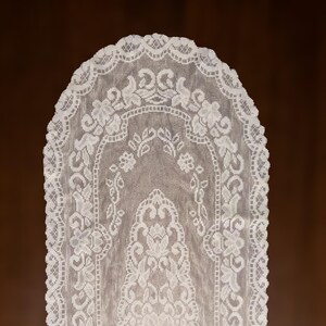 May include: A white lace table runner with a floral and leaf pattern. The runner has a scalloped edge and is displayed against a brown background. The delicate design is visible throughout the fabric.