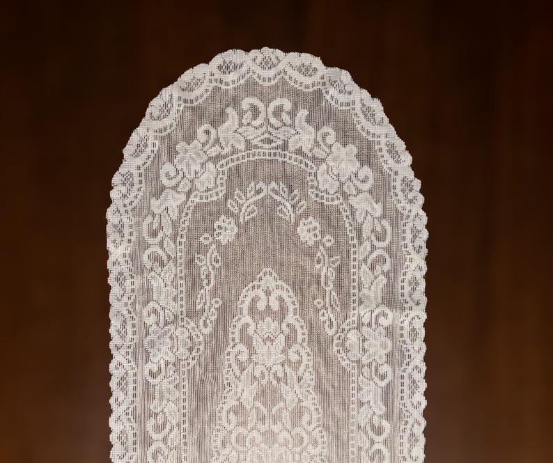 May include: A white lace table runner with a floral and leaf pattern. The runner has a scalloped edge and is displayed against a brown background. The delicate design is visible throughout the fabric.