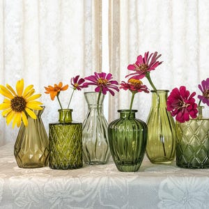 May include: A collection of six small glass vases in various shades of green, each featuring unique textures and shapes. The vases are filled with vibrant, colorful flowers including a large yellow sunflower and several bright pink and orange zinnias.