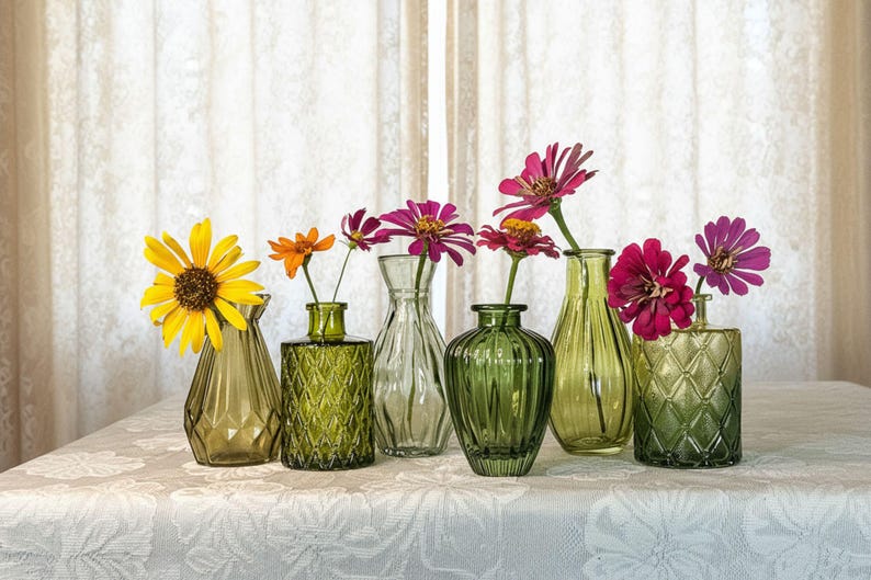 May include: A collection of six small glass vases in various shades of green, each featuring unique textures and shapes. The vases are filled with vibrant, colorful flowers including a large yellow sunflower and several bright pink and orange zinnias.