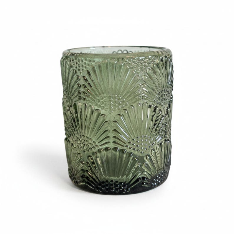 K&ouml;nnte beinhalten: Ein zylindrischer Kerzenhalter aus Glas in einem ged&auml;mpften Olivgr&uuml;n mit einem sich wiederholenden Pr&auml;gemuster aus stilisierten f&auml;cherf&ouml;rmigen Bl&uuml;tenbl&auml;ttern und kleinen kreisf&ouml;rmigen Punkten, die eine strukturierte Oberfl&auml;che bilden.