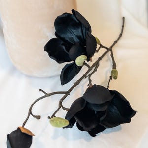 May include: A sprig of artificial black magnolia flowers with green buds and a brown stem. The flowers have multiple petals and are arranged in a naturalistic way. The background is a soft, neutral color.