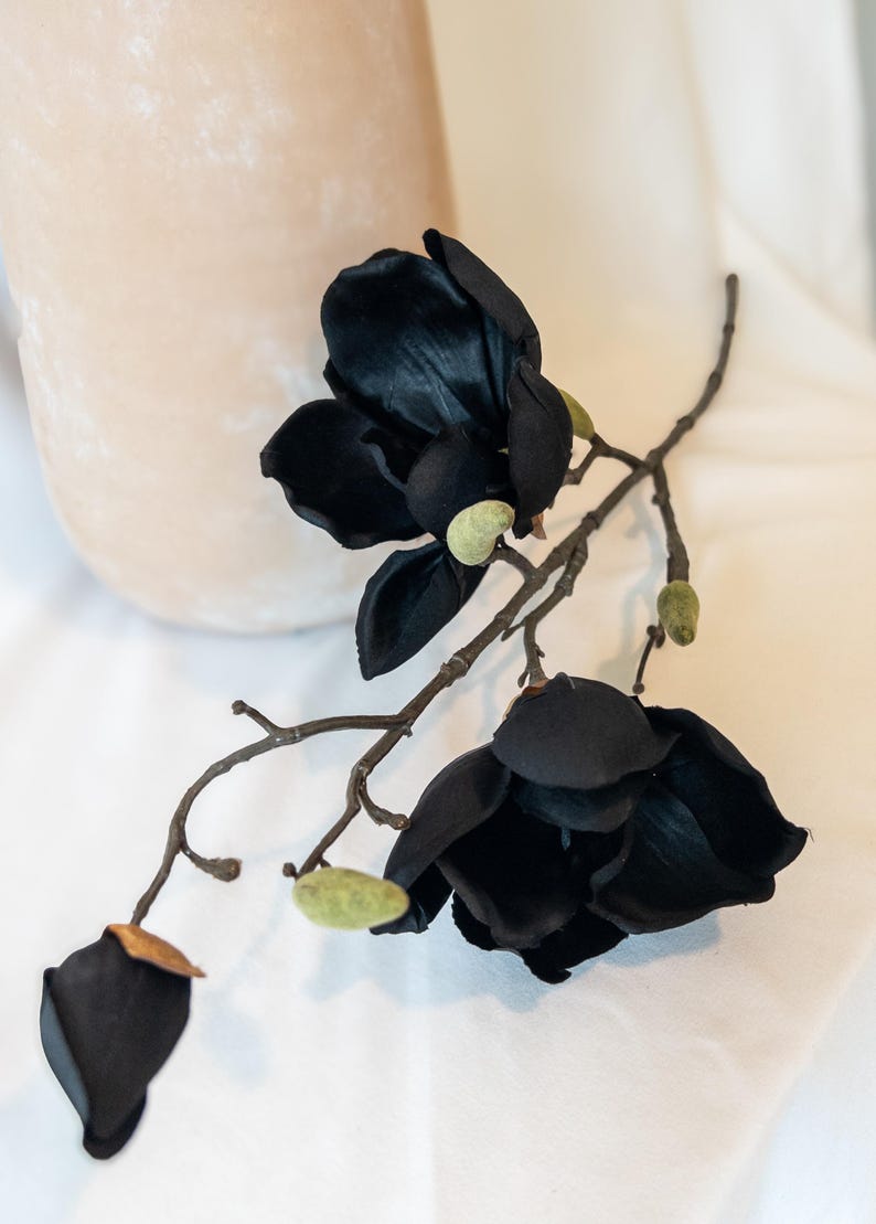 May include: A sprig of artificial black magnolia flowers with green buds and a brown stem. The flowers have multiple petals and are arranged in a naturalistic way. The background is a soft, neutral color.