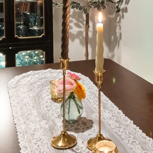 May include: A table setting featuring two lit candles in gold candlesticks, a small vase with flowers, and a gold votive candle holder. A white lace runner decorates the dark wooden table. A cabinet with dishes is in the background.