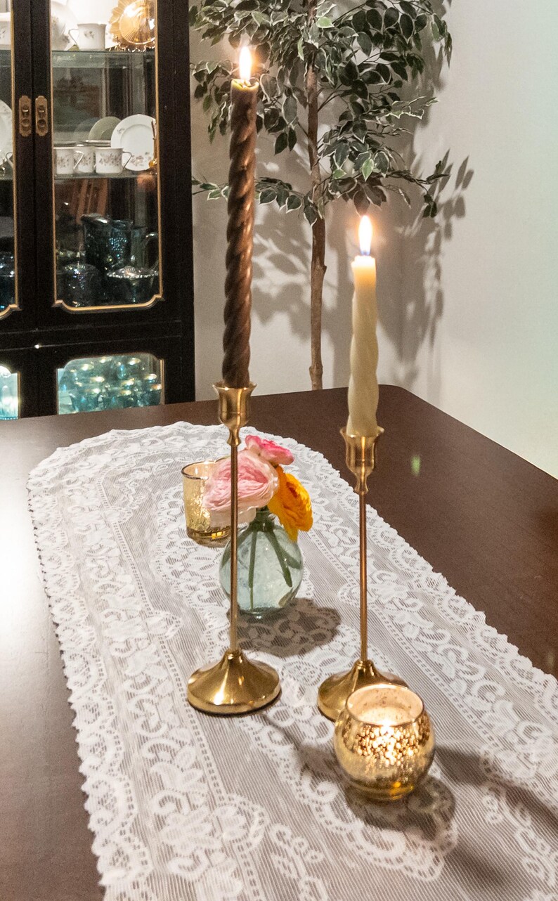 May include: A table setting featuring two lit candles in gold candlesticks, a small vase with flowers, and a gold votive candle holder. A white lace runner decorates the dark wooden table. A cabinet with dishes is in the background.