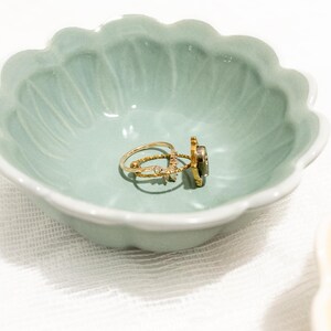 May include: A light green, scalloped-edge ceramic dish holds three gold rings. The rings have various designs, including a band with small stones and a ring with a dark stone. The dish is on a white surface.