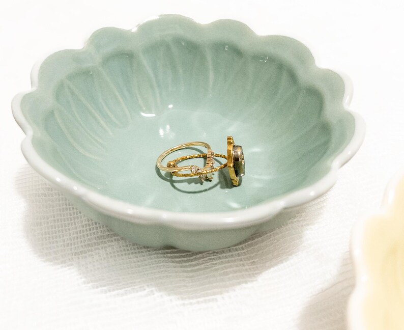 May include: A light green, scalloped-edge ceramic dish holds three gold rings. The rings have various designs, including a band with small stones and a ring with a dark stone. The dish is on a white surface.